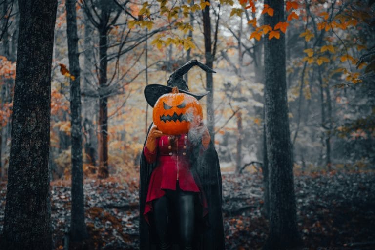 A witch in the woods holding up a pumpkin to cover her face